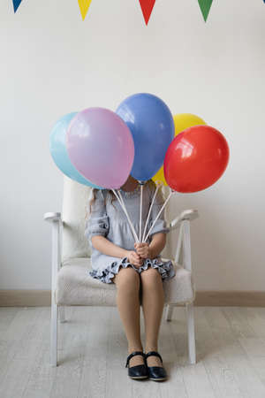 Cute little girl on a birthday sitting on a white armchair in a room with an armful of multi-colored balloons. She hid herself. The face is not visible. The atmosphere of the holiday. Happiness for a young lady. Home celebration concept. White background. Clean and bright apartments.の写真素材