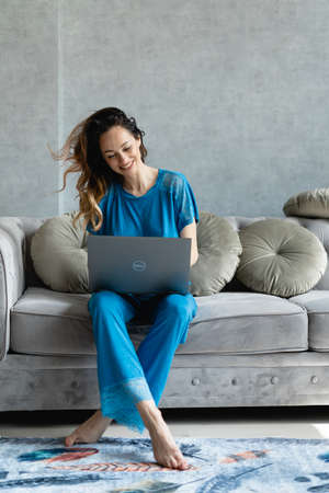 Cool young woman in blue pajamas is sitting on the couch. A laptop is kneeling and legs crossed on the floor. Webcam chat. Conference call with business partners from home. Video chat on the day off. The concept of the use of technology in business development.の写真素材