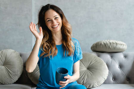 Cute sexy girl looking at the web camera while sitting on a sofa with a cup of espresso in her hands. She sits on the couch and waves hello to her conferencing partner. Home affairs business lady. Video communication in the life of modern youth.の写真素材