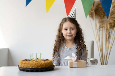 A beautiful girl of 6-7 years celebrates her birthday. She is sitting and a table is standing in front of her. It has a birthday cake and a small box with a gift. The room is decorated for the holiday. It is light and comfortable. Holiday concept at home.の写真素材