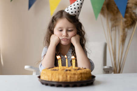 Cute baby on his birthday sits at the table propping his head with two fists. In front of her is a cake with candles. She has a birthday and no mood. The child is upset and sad. Bright room and holiday atmosphere.の写真素材