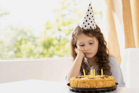 Cute baby sitting at the table. Girls birthday and birthday cake with candles. The child is sad and laid his head on his raised hand. In the background is a large window. The room is bright and comfortable. Place for an inscription. Holiday concept for daughter.の写真素材