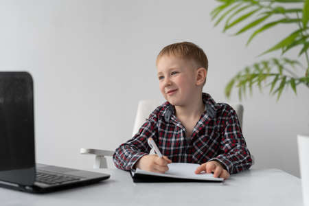 A schoolboy is doing quarantine homework at home. Distance learning and lessons. The boy sits on a chair at the table. The task in the laptop. Online education and quarantine. Place for text.の写真素材
