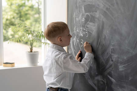 A modern student in the classroom writes math examples on the blackboard. He stands at the side of the window. Math lesson in elementary school. Hands of a boy lie on a blackboard. Place for text.の写真素材