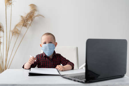 Home schooling education. Online education. A boy sits and does homework at a laptop. He has a pen in his hands and there is a notebook on the table. A medical mask is on the face. Distance education of children. Place for text.の写真素材