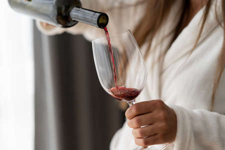 Close up of a bottle in female hands. Pour red wine into a glass. Woman in a hotel room. Bathrobe.の写真素材