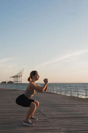 the girl is engaged in fitness by the sea or ocean, crouches and smiles, in the morning light the concept of an active lifestyle. Place for textの写真素材