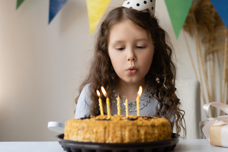 Sad girl blows out the candles on the holiday cake. She is at home alone in a bright room decorated...