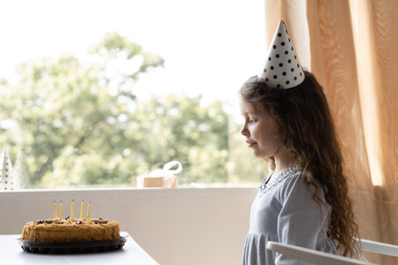 Cute girl with her long curly hair is sitting sideways near the window. Beautiful baby profile and...