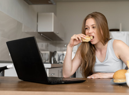 Young pretty caucasian woman enjoying her snack in kitchen by the laptop while working remotely from...