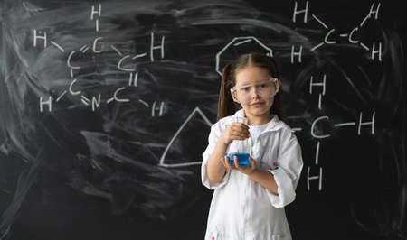 little girl schoolgirl in goggles and a white coat stands near the chalkboard. Chemical formulas. Hold a test tube with liquid in your hands. Back to school concept.の写真素材