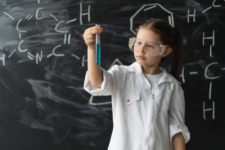 little girl schoolgirl in goggles and a white coat stands near the chalkboard. lifts the test tube...