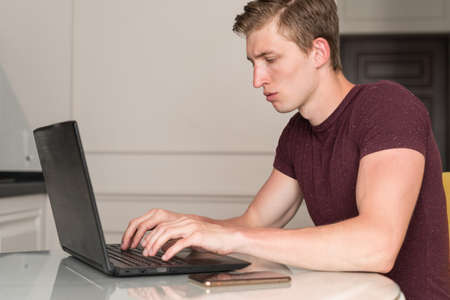 Focused serious young man in t-shirt sitting at home in the kitchen, working on laptop computer. Remote job, technologies conceptの写真素材