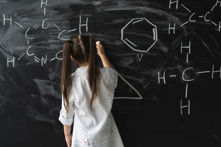 Cute little girl writing on a blackboard at a chemistry lesson in class. White coat and goggles. Place for textの写真素材