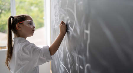 little girl schoolgirl near the chalkboard solves a chemistry problem. White coat and goggles. Offline education conceptの写真素材