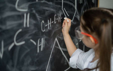 little girl schoolgirl near the chalkboard solves a chemistry problem. Write formulas for elements. White coat and goggles. Offline education conceptの写真素材