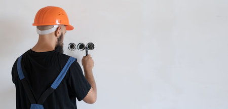 a male electrician wearing blue work clothes and an orange hard hat. Stands near the wall looking at...