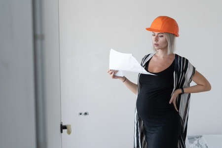 young pregnant woman wearing a protective construction helmet on her head. Looks at the task sheet. Stands against a white wall.の写真素材