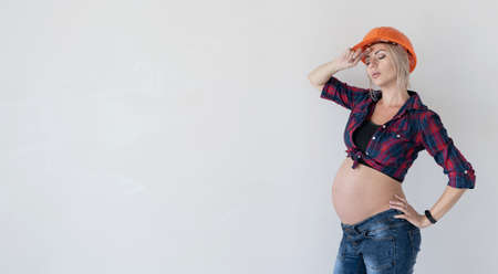 Young beautiful pregnant blonde woman. protective construction mask on the head. He wipes sweat from his forehead with his hand. Free space for textの写真素材