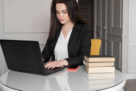 A portrait of a designer working at home on new ideas. a dark-haired woman sits at a white table in formal clothes, looks into a laptop. Stack of books nearbyの写真素材
