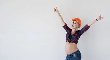 young beautiful pregnant woman wearing a red plaid shirt and a construction safety helmet. spread her arms to the sides, rejoices. Place for textの写真素材
