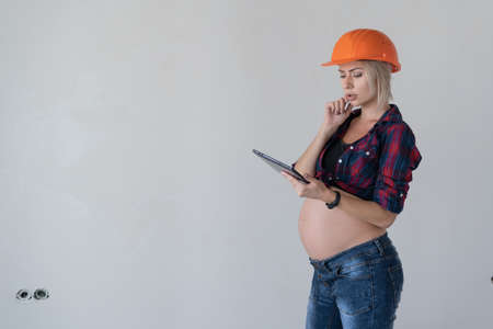 young beautiful pregnant blonde woman stands near the wall. Working womens clothing shirt in a cage orange construction helmet. Hold the tablet in hand to read information and think. Copy space.の写真素材