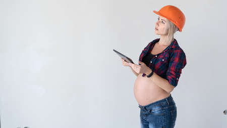 young beautiful pregnant blonde woman stands near the wall. Work women's clothing shirt in a cage orange construction safety helmet. hold the tablet in your hands, look away to the free spaceの写真素材
