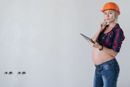 young beautiful pregnant blonde woman stands near the wall. Working womens clothing shirt in a cage orange construction helmet. Hold the tablet in your hands and look at the camera. Copy space.の写真素材