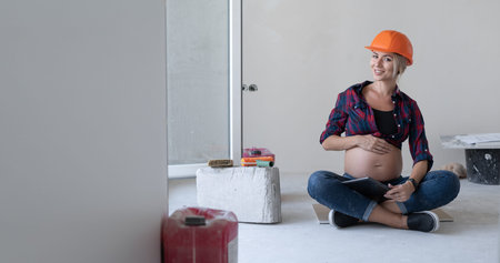 pregnant blonde woman sits on the floor in the room where renovations are in progress. a protective helmet is worn over the head. hand stroking the belly. looks at the photographer. Copy space.の写真素材