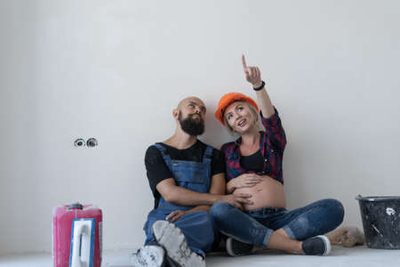 husband and his pregnant wife are sitting on the floor in the room. work uniform and orange safety helmet. point up and look at free space for text.の写真素材