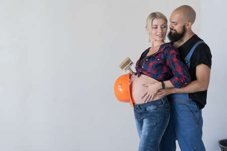 A man hugs his pregnant wife. work uniform and orange safety helmet. happy family makes renovation. Copy space.の写真素材