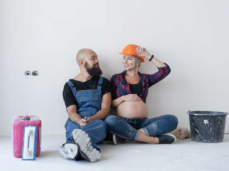 husband and his pregnant wife are sitting on the floor in the room. work uniform and orange safety helmet. look at each other and smile. Copy space.の写真素材