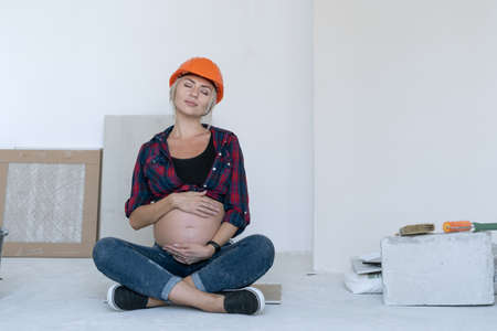 pregnant blonde woman sits on the floor in the room where renovations are in progress. a protective helmet is worn over the head. strokes her stomach with both hands, closed her eyes and enjoys the momentの写真素材