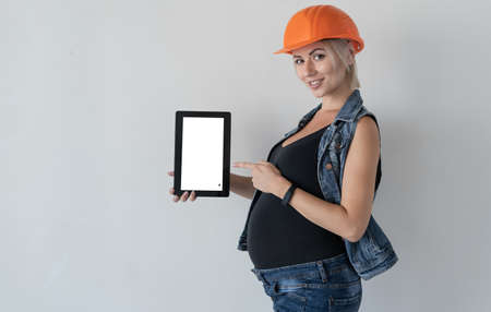 Young beautiful woman builder in an orange protective helmet on her head. Pregnant girl. Stands near the wall holding a tablet in his hand. White screen. Point your hand. Home renovation concept before childbirth.の写真素材