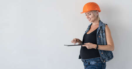 Young beautiful woman builder in an orange protective helmet on her head. Pregnant girl. Stands near the wall holding a tablet in his hand, typing a message. Home renovation concept before childbirth.の写真素材