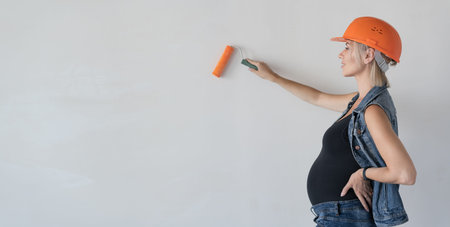 Young beautiful woman builder in an orange protective helmet on her head. Pregnant girl. Paints the wall with a paint roller. Place for textの写真素材