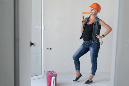 Young beautiful woman builder in an orange protective helmet on her head. Pregnant girl. stands indoors with a paint roller in his hands. Place for textの写真素材
