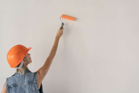 Young beautiful woman builder in an orange protective helmet on her head. Pregnant girl. Paints the wall with a paint roller. Close-up of a hand and a tool. Place for textの写真素材