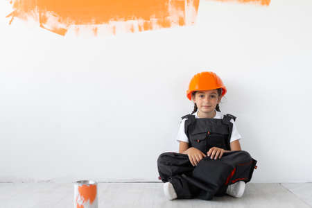the girl is dressed in overalls and an orange protective helmet on her head. sitting on the floor looking in front. Place for text.の写真素材