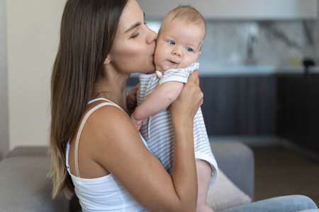 young beautiful mom is sitting on the couch. Holds a newborn baby in her arms. kiss a child on the...