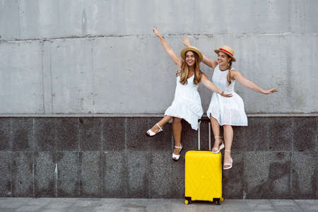 Beautiful two female tourist twins are sitting near a yellow suitcase with their arms spread out to the sides imitate flight. Flying an airplane conceptの写真素材