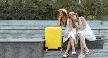 tired two female twin tourists are sitting near a yellow suitcase on the steps. Flight cancellation long wait. Copy spaceの写真素材