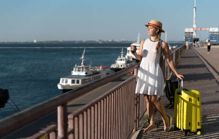 A girl traveler in a dress and hat stands on the pier and looks at the ships in the sea. She is holding a yellow suitcase with things and she has emotions of joy from the trip.の写真素材