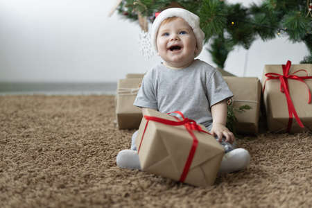 a little boy in a Santa hat sits on the floor near the Christmas tree and holds a box with a gift in his hands. Christmas winter holiday conceptの写真素材