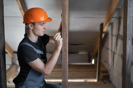 male builder dressed in work clothes and an orange hard hat. Measures the distance with a tape measure and puts a mark on the board. Copy spaceの写真素材