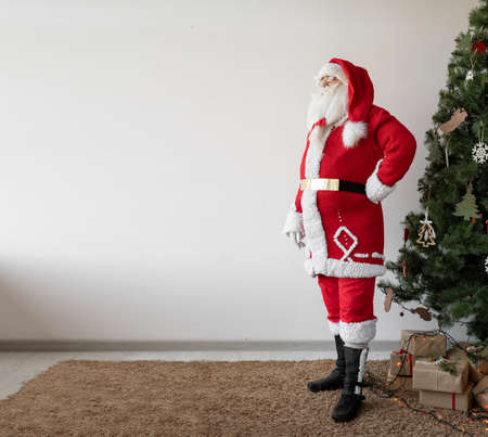 Santa Claus stands near the Christmas tree with his hands on his hips. Looks at empty space for textの写真素材