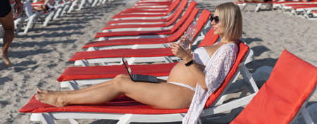 pregnant woman dressed in a white swimsuit lies on a sun lounger, working on a laptop copy spaceの写真素材