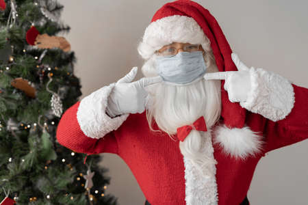 Santa Claus is standing near the Christmas tree, his index fingers point to a medical mask on his face. Copy spaceの写真素材