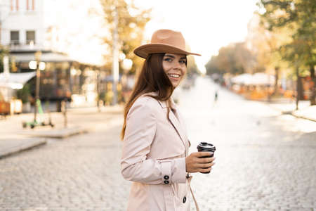 young woman walks through the city streets dressed in a hat and a raincoat. Coffee to go. Copy spaceの写真素材