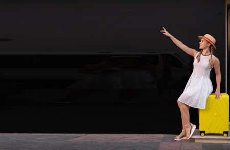 A girl traveler with a yellow suitcase in her hand stands on the street near a black background. point with your index finger to an empty space for text.の写真素材